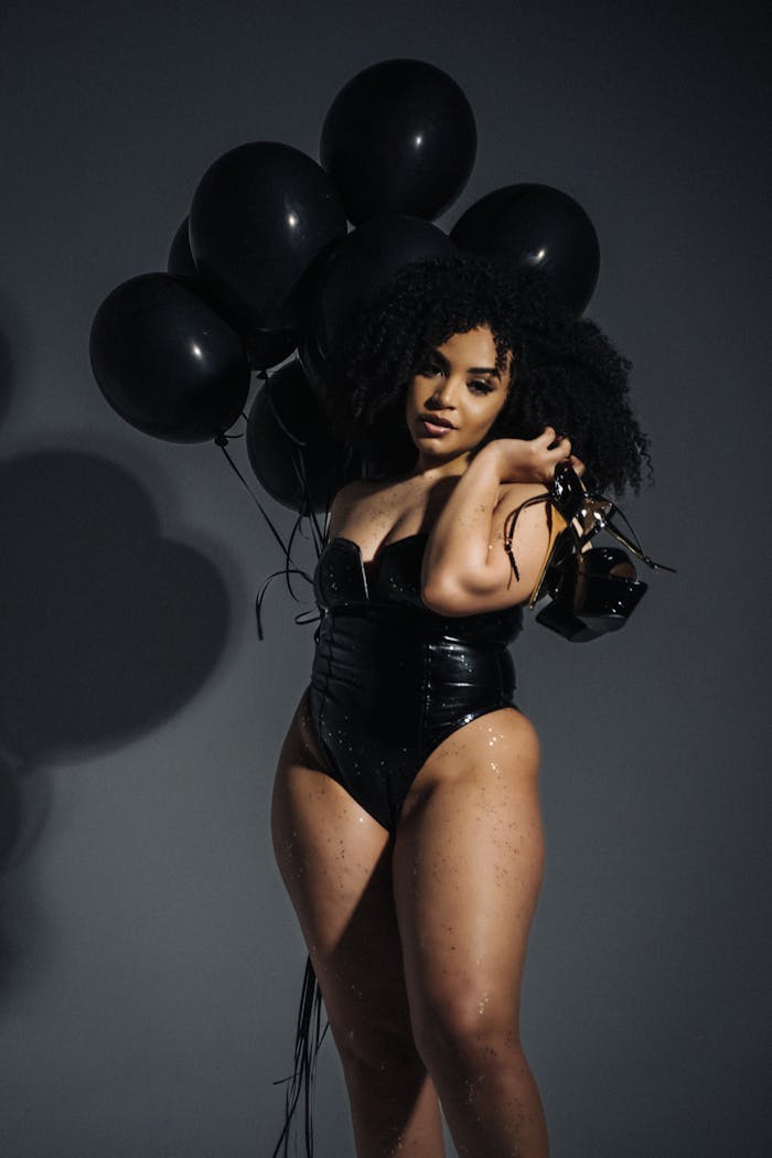 menu-08 Fashionable woman in a black latex bodysuit poses in a studio with black balloons against a gray background.